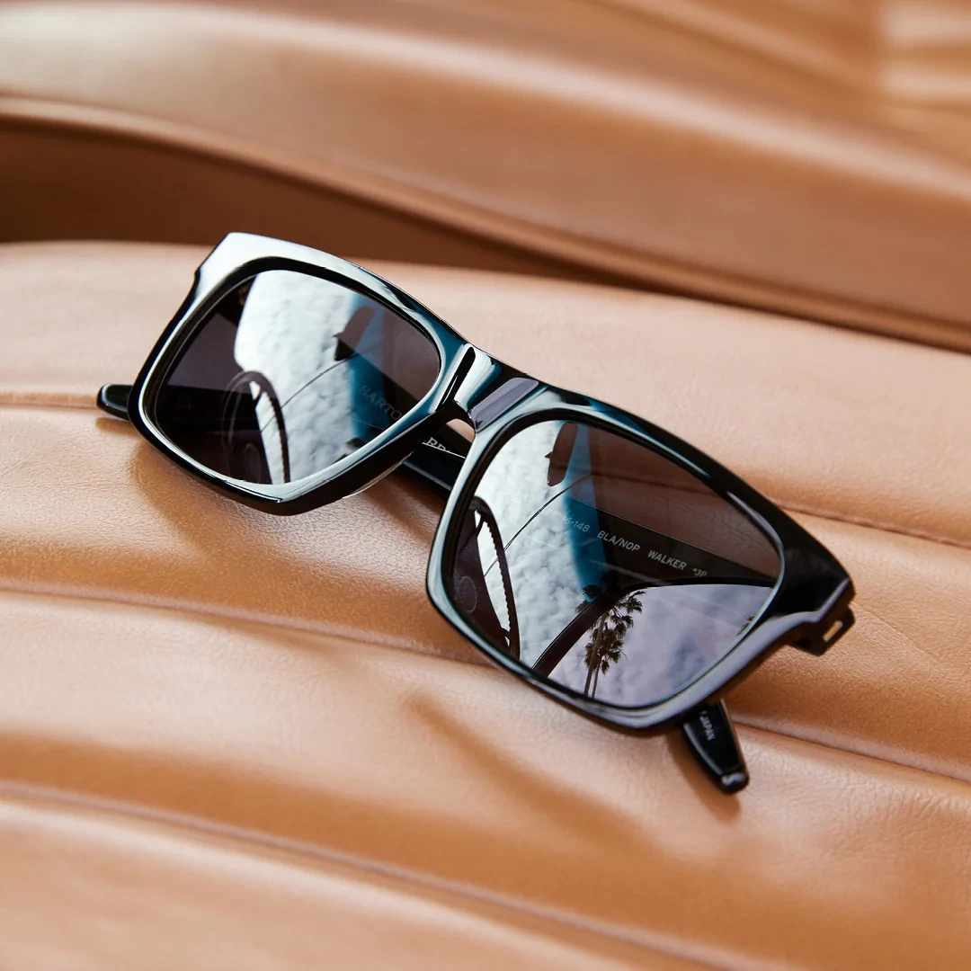 Lunettes de soleil Barton Perreira noires rectangulaires posées sur un siège en cuir fauve